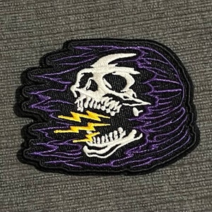 Könnte beinhalten: Ein gestickter Patch mit einem schwarzen und weißen Totenkopf, aus dessen Mund ein Blitzschlag kommt. Der Totenkopf ist von einem violetten und schwarzen Wirbelmuster umgeben.