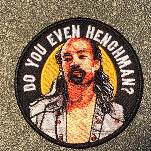 Pode incluir: Patch bordado com o texto "DO YOU EVEN HENCHMAN?" em letras brancas sobre fundo preto. O patch apresenta um retrato de um homem com cabelo comprido e bigode, sobre um fundo amarelo e laranja.