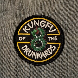 May include: A round black patch with the words "KUNGFU OF THE DRUNKARDS" in white lettering. A green and red snake-like figure is in the center. The patch has a yellow border and is on a gray denim background.