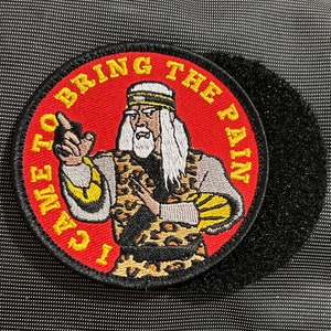 Pode incluir: Etiqueta bordada vermelha e amarela com o texto "I Came To Bring The Pain" e uma imagem de desenho animado de um homem com barba e cabelo comprido vestindo uma camisa de estampa de leopardo.