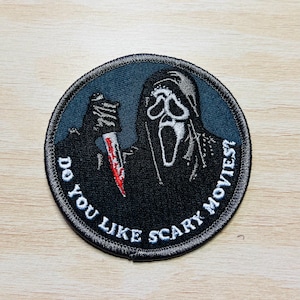 May include: A black and white embroidered patch featuring a ghost-like figure holding a knife. The text surrounding the figure reads "Do you like scary movies?"