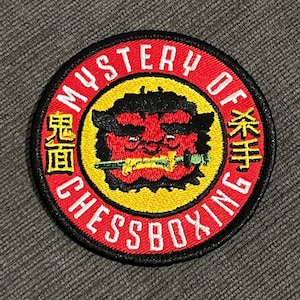 May include: Circular embroidered patch with a red border and the words "MYSTERY OF CHESSBOXING" in white. The patch features a red and black cartoon demon face with a yellow background and Japanese text.