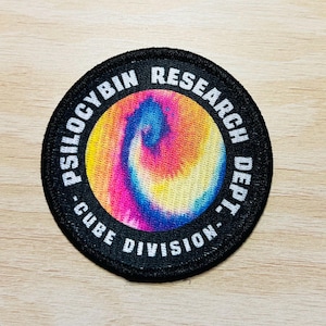 Può includere: Toppa ricamata in bianco e nero con un design a spirale psichedelico in rosa, giallo, blu e arancione. Il testo "PSILOCYBIN RESEARCH DEPT. - CUBE DIVISION" è scritto in un cerchio attorno alla spirale.