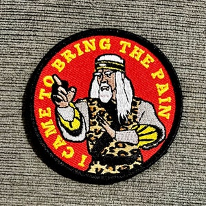 Pode incluir: Um remendo redondo com borda preta e fundo vermelho. O remendo apresenta uma imagem de desenho animado de um homem com barba branca e cabelo branco comprido usando uma túnica de estampa de leopardo e uma faixa amarela. O texto "I Came To Bring The Pain" está escrito em amarelo ao redor da borda do remendo.