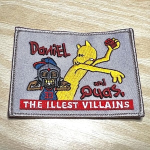 May include: A grey embroidered patch with the text "Daniel and Quas, The Illest Villains". The patch features a cartoon image of a yellow character with a red brick in its hand and a black character wearing a helmet and a red jersey with the number 33.