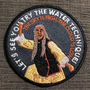 May include: A round, blue embroidered patch with a white outline. The patch features a cartoon image of a man with long white hair and a beard, holding a stick. The text on the patch reads "Let's see you try the water technique. The sky is high, the cloud is low."