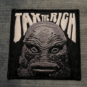 Peut inclure: Écusson carré noir avec le texte blanc « TAX THE RICH » au-dessus d'une illustration grise détaillée d'un visage de créature. Le patch est destiné à être appliqué sur des vêtements ou des accessoires.