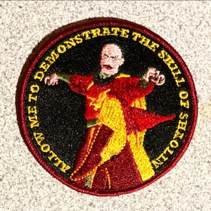 May include: Embroidered patch featuring a cartoon character in a red and yellow costume with the text "Allow me to demonstrate the skill of shadows".