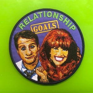 May include: Embroidered patch with a purple background and the text "Relationship Goals" in green. The patch features a cartoon image of a man and woman with a yellow box with the text "Goals" in the center.