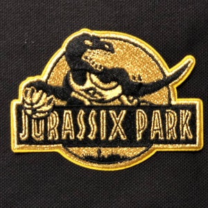 Puede incluir: Parche bordado con un diseño de dinosaurio negro y detalles dorados, sobre un fondo circular dorado. El texto "JURASSIC PARK" está en letras doradas dentro de un borde rectangular negro. El parche está sobre tela negra.