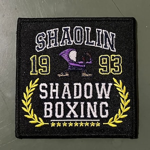 Peut inclure: Écusson brodé noir avec des lettres jaunes et blanches. L'écusson porte l'inscription "Shaolin 1993 Shadow Boxing" avec une figure violette et blanche au centre.