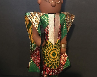 May include: A wooden doll wearing a green, yellow, and red patterned robe with a gold crown and glasses. The doll is holding a black walking stick and is standing on a green base.