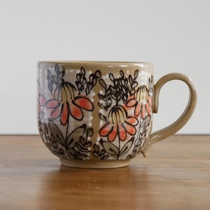 May include: A ceramic mug with a light tan base color featuring a hand-painted floral pattern. The design includes orange and yellow flowers with dark brown stems and leaves, accented by vertical white dotted lines around the exterior surface.