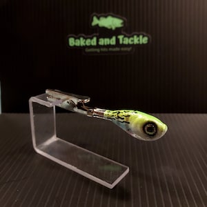 May include: A green and white fishing lure with black accents, held by a silver clip on a clear acrylic stand. The lure has a detailed eye and is designed to resemble a small fish. The background features the text "Baked and Tackle".
