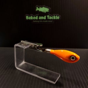 May include: An orange and black fishing lure with a silver clip, displayed on a clear acrylic stand. The lure has a detailed eye and is set against a black background with the text "Baked and Tackle".