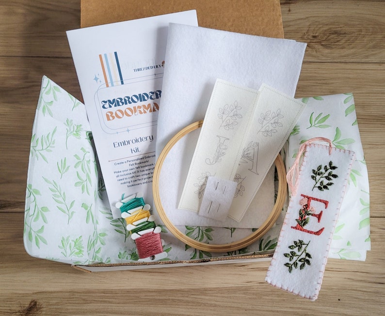 May include: Embroidery kit for making personalized bookmarks. The kit includes a white felt bookmark with a pre-printed design of a letter E and leaves, a wooden embroidery hoop, white felt, two pre-printed designs for bookmarks, embroidery floss, and instructions.