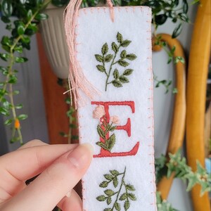 May include: White felt bookmark with pink stitching and a pink tassel. The bookmark features an embroidered red letter "E" with green leaves and pink flowers around it. There are two green leaf sprigs above and below the letter.