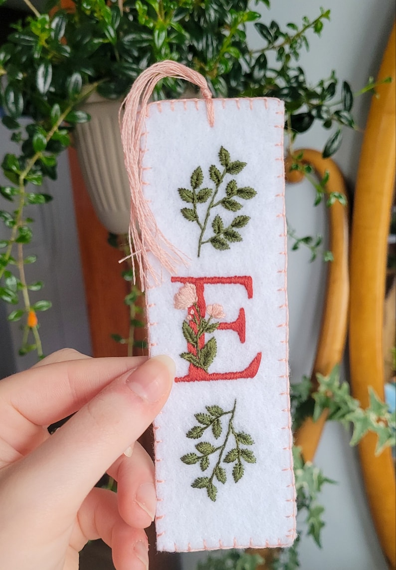 May include: White felt bookmark with pink stitching and a pink tassel. The bookmark features an embroidered red letter "E" with green leaves and pink flowers around it. There are two green leaf sprigs above and below the letter.