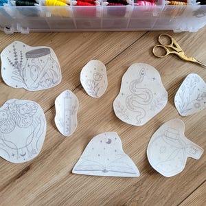 May include: Eight black and white line drawings on white paper, featuring a moth, a snake, a book, a jar, a vial, and flowers. The drawings are all in a whimsical style and would be perfect for embroidery or other crafts.