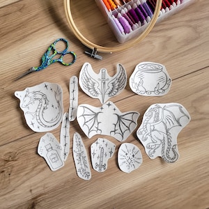 Könnte beinhalten: Ein Set mit 10 Bügelbildern für Stickereien mit verschiedenen Designs, darunter ein Drache, ein Kessel, ein Schwert und eine Flasche. Die Muster sind auf weißem Papier gedruckt und bereit zum Aufbügeln auf Stoff.