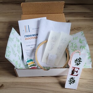 May include: A white box with a green and white patterned tissue paper lining. The box contains a felt embroidery kit with a hoop, embroidery floss, needles, and two felt bookmarks with embroidery patterns. The box also includes instructions for creating a personalized felt bookmark.