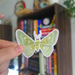 May include: A green and white embroidered moth sticker with detailed wing patterns. The sticker features a white border and is held in front of a blurred background of a bookshelf.