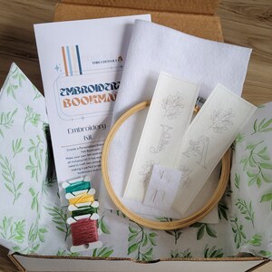 May include: A craft kit for making embroidered felt bookmarks. The kit includes two pre-printed felt bookmarks, embroidery floss, a wooden embroidery hoop, and needles. The kit is packaged in a brown cardboard box with a white tissue paper liner.