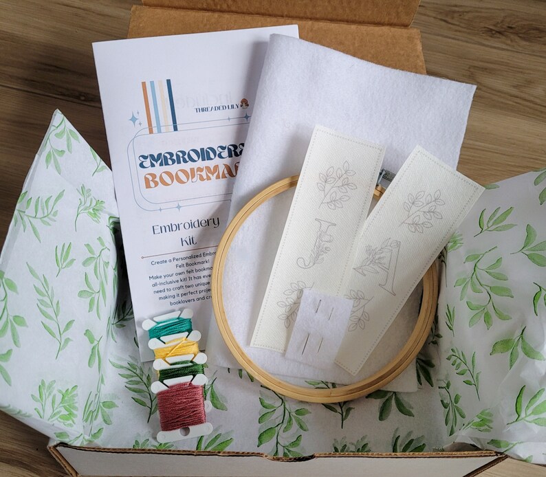 May include: A craft kit for making embroidered felt bookmarks. The kit includes two pre-printed felt bookmarks, embroidery floss, a wooden embroidery hoop, and needles. The kit is packaged in a brown cardboard box with a white tissue paper liner.