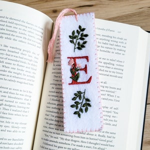 May include: A white felt bookmark with a pink tassel and embroidered letter E in red. The letter is surrounded by green leaves and pink flowers. The bookmark is on a page of a book with text about faeries.