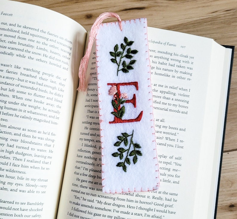 May include: A white felt bookmark with a pink tassel and embroidered letter E in red. The letter is surrounded by green leaves and pink flowers. The bookmark is on a page of a book with text about faeries.
