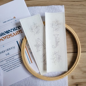 May include: Two white fabric bookmark patterns with embroidery designs of leaves and letters. The patterns are on a white surface with a wooden embroidery hoop.