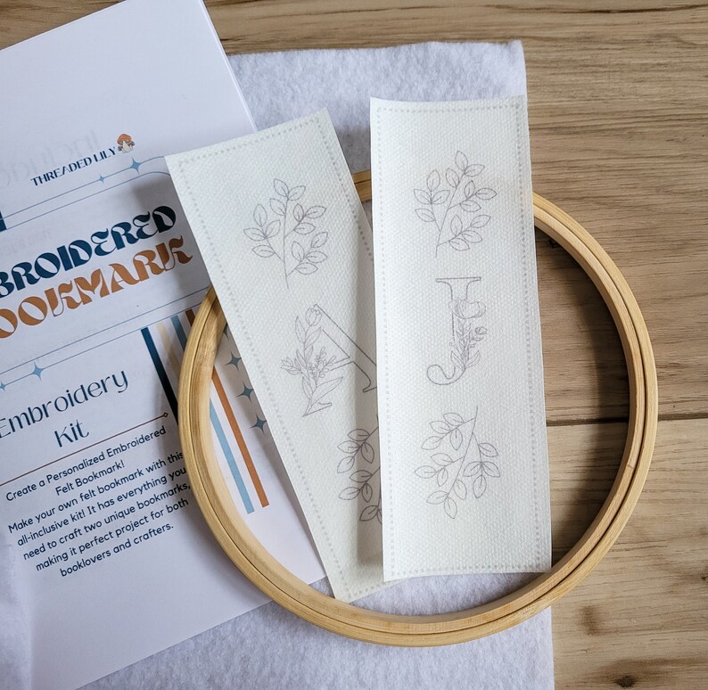 May include: Two white fabric bookmark patterns with embroidery designs of leaves and letters. The patterns are on a white surface with a wooden embroidery hoop.