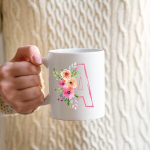 May include: White ceramic mug featuring a pink floral design and the letter "A" in pink. The mug is being held by a person's hand.