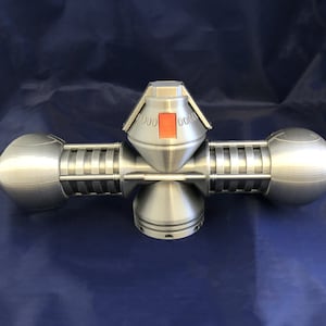 May include: A silver 3D printed model of a futuristic spaceship with a red button on the top. The model has a cylindrical body with two rounded ends and a central dome with a red button.