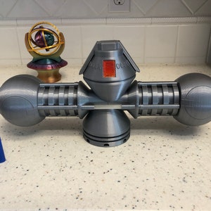 Puede incluir: Una escultura geométrica gris, impresa en 3D, con una estructura central y cuatro extensiones esféricas. La escultura tiene un diseño detallado con un acento rojo. Un encendedor azul está en primer plano.