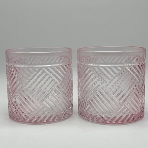 May include: Two pink-tinted, clear glass tumblers. Each tumbler has a cylindrical shape with a thick, flat base and a straight rim. The exterior features a crisscross pattern, creating a textured effect. The glasses are empty and sit side-by-side.