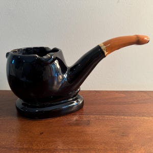 May include: Black ceramic pipe ashtray with a brown and gold handle. The ashtray is shaped like a pipe and has a round base.