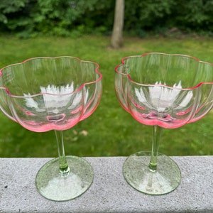 May include: Two clear glass stemmed drinking glasses with a pink rim. The glasses have a scalloped edge and a wide bowl.