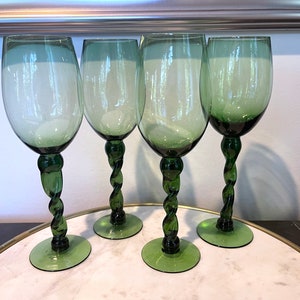May include: Four green-tinted wine glasses with a unique twisted stem design. The glasses have a classic goblet shape and sit on a round base. The glasses are displayed on a white and gold tray.