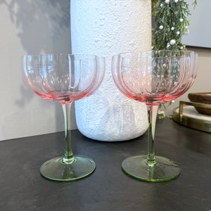 May include: Two pink stemmed glasses with a fluted design. The glasses have a green base and are sitting on a dark surface.