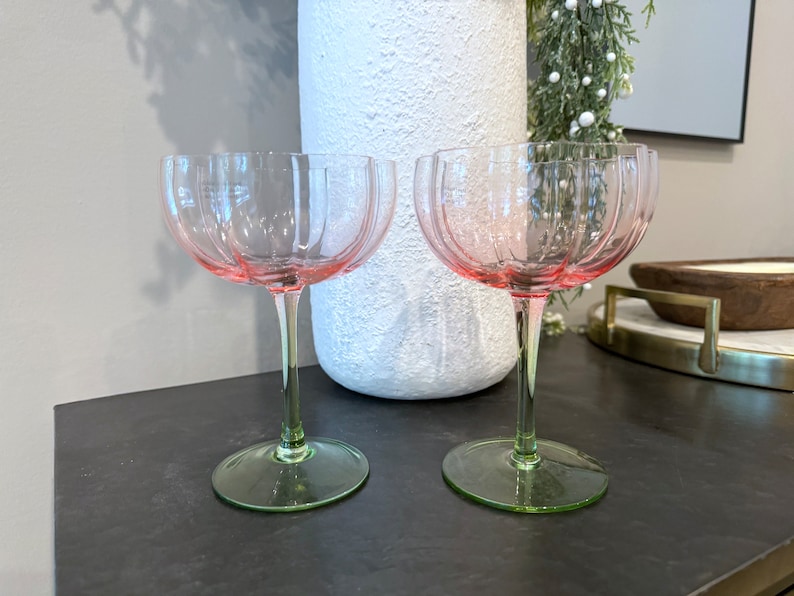 May include: Two pink stemmed glasses with a fluted design. The glasses have a green base and are sitting on a dark surface.