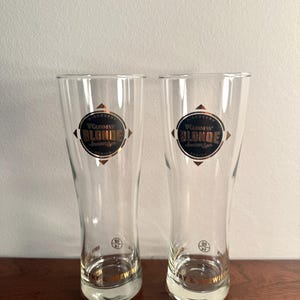 Puede incluir: Dos vasos de cerveza de vidrio transparente con borde dorado y las palabras "Guinness Blonde American Lager" impresas en la parte delantera. Los vasos est&aacute;n sobre una superficie de madera.