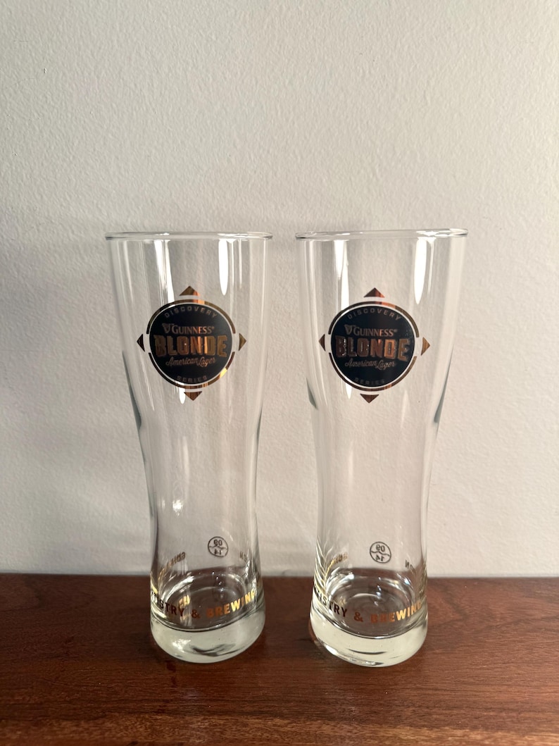 Puede incluir: Dos vasos de cerveza de vidrio transparente con borde dorado y las palabras "Guinness Blonde American Lager" impresas en la parte delantera. Los vasos est&aacute;n sobre una superficie de madera.