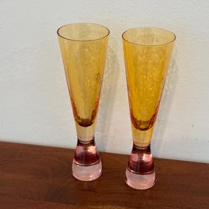 May include: Two champagne flutes with textured, amber-colored glass. The stems are a translucent pink. The glasses are displayed on a dark brown wooden surface against a white wall.