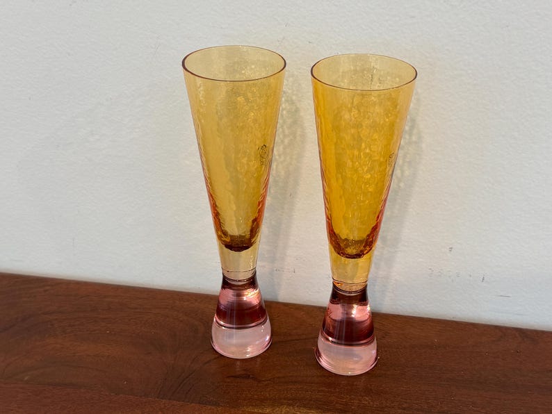 May include: Two champagne flutes with textured, amber-colored glass. The stems are a translucent pink. The glasses are displayed on a dark brown wooden surface against a white wall.