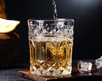 Può includere: Un bicchiere di vetro trasparente con un motivo a forma di diamante è riempito di un liquido dorato. Il bicchiere è appoggiato su un sottobicchiere di legno.