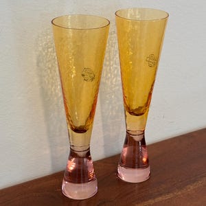 May include: Two amber-colored champagne flutes with textured glass. The stems are a soft pink, and each glass has a small, circular emblem near the top. The flutes are displayed on a wooden surface.