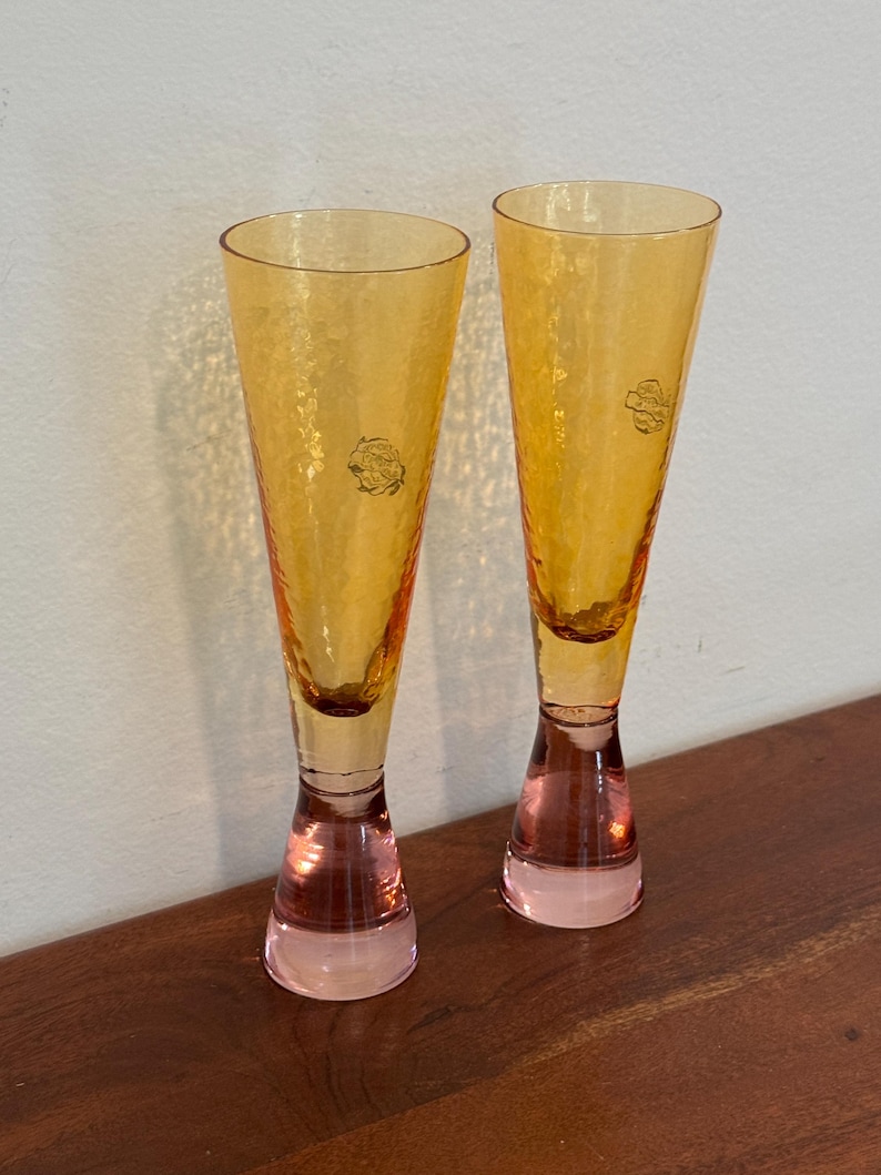May include: Two amber-colored champagne flutes with textured glass. The stems are a soft pink, and each glass has a small, circular emblem near the top. The flutes are displayed on a wooden surface.