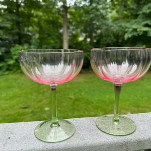 May include: Two clear glass champagne coupes with pink rims and green stems. The glasses are sitting on a wooden surface.