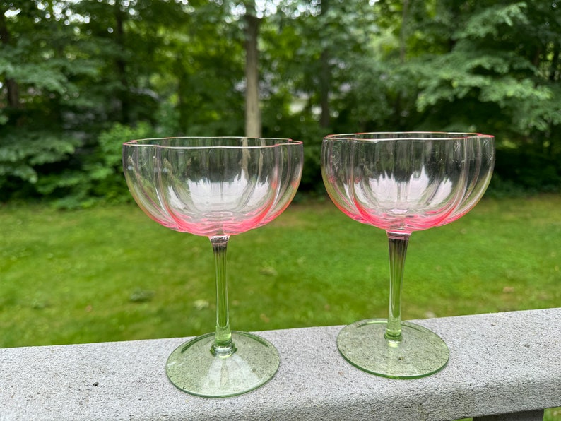 May include: Two clear glass champagne coupes with pink rims and green stems. The glasses are sitting on a wooden surface.